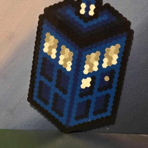 May include: A blue, black, and white pixel art representation of a police box. The box has a dark blue body with lighter blue panels and white windows. The edges are outlined in black, and the top features a small white detail.