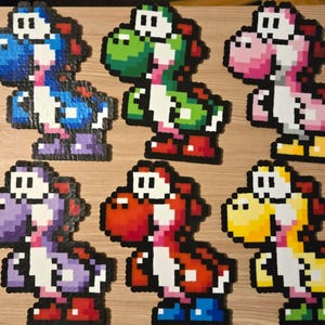 May include: Six colorful pixel art figures of Yoshi, a dinosaur character from the Super Mario video game series. Each figure is a different color: blue, green, pink, purple, red, and yellow. The figures are made of small, square beads.