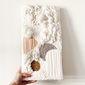 May include: A rectangular, textured wall hanging with a white background and various raised, abstract shapes. The design includes peach, brown, and gold elements, with a fluffy, cloud-like texture. The piece is held by a hand.