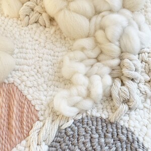 May include: Close-up of a textured fiber art piece. The artwork features a combination of woven and knotted textures in shades of white, cream, light gray, and a touch of blush pink. The piece has a soft, tactile appearance.