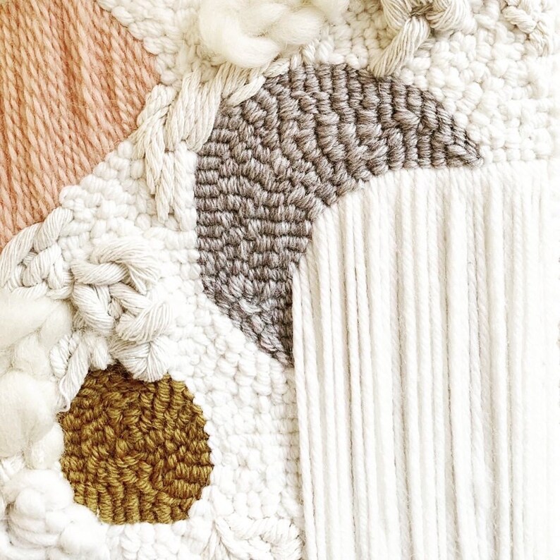 May include: Close-up of a woven wall hanging with a textured design. The piece features various textures and colors, including peach, white, gray, and gold. The design incorporates knotted and braided elements, creating a tactile and visually interesting artwork.