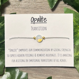 May include: Opalite bracelet on a card. The card reads "Opalite Transition" and includes a description of the stone's properties. The bracelet features a small, round opalite bead with gold accents on a gray cord.