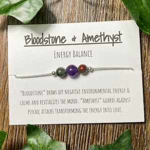May include: A bracelet featuring three gemstone beads: green, purple, and red. The card reads "Bloodstone & Amethyst, Energy Balance." The text below describes the properties of the stones. The bracelet is strung on a gray cord.