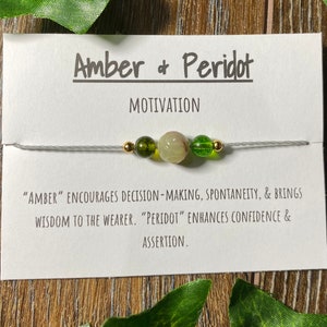 May include: A white card with the text "Amber & Peridot" and "MOTIVATION" in black. The card also has the text "Amber" encourages decision-making, spontaneity, & brings wisdom to the wearer. "Peridot" enhances confidence & assertion."  A string bracelet with green and gold beads is displayed on the card.