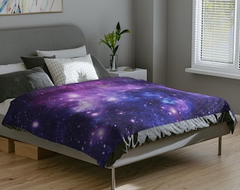 Puede incluir: Una manta suave con un diseño de galaxia vibrante en tonos morados, azules y blancos. La manta está colocada sobre una cama, mostrando su textura suave y su patrón celestial. La manta es una adición acogedora a cualquier dormitorio.