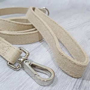 Puede incluir: Una correa de perro de lona beige con un clip de metal plateado. La correa está hecha de un material grueso y duradero, ideal para pasear a tu perro.