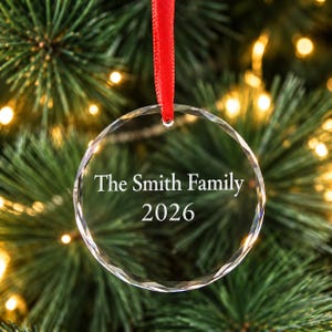 Personalised family Christmas ornament: clear engraved glass bauble reading The Smith Family 2026 with red ribbon, hanging on a lit Christmas tree