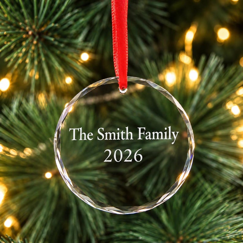Personalised family Christmas ornament: clear engraved glass bauble reading The Smith Family 2026 with red ribbon, hanging on a lit Christmas tree