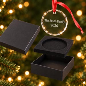 Engraved glass family Christmas bauble reading The Smith Family 2026 with red ribbon above an open black gift box, free gift box included