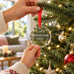 Hands hanging a personalised engraved glass family Christmas bauble with red ribbon on a decorated Christmas tree in a cosy living room