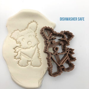 May include: A brown plastic cookie cutter in the shape of a Yorkshire Terrier dog. The cutter is shown next to a white impression of the cutter in a block of modeling clay. The text "DISHWASHER SAFE" is printed in blue on a white background.