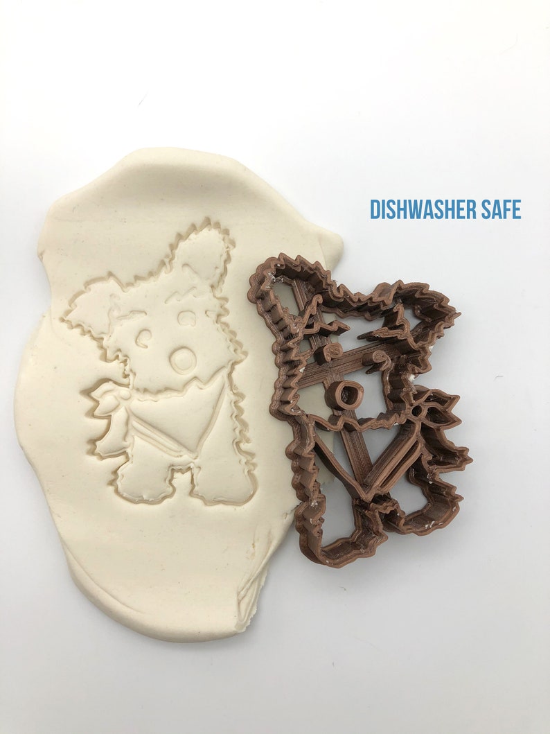 May include: A brown plastic cookie cutter in the shape of a Yorkshire Terrier dog. The cutter is shown next to a white impression of the cutter in a block of modeling clay. The text "DISHWASHER SAFE" is printed in blue on a white background.