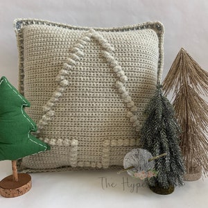 Peut inclure: Un coussin crocheté avec un fond beige et un motif d'arbre blanc. Le coussin est entouré de trois petits sapins de Noël artificiels, un vert, un argenté et un brun.