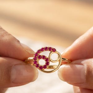 Może przedstawiać: Złoty pierścionek z symbolem zodiakalnego Raka. Lewa strona symbolu jest wysadzana małymi, okrągłymi, żywymi różowo-czerwonymi kamieniami szlachetnymi, a prawa strona składa się z gładkiej, polerowanej, okrągłej złotej pętli.
