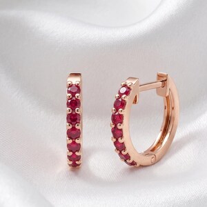 Könnte beinhalten: Ein Paar roségoldene Creolen. Jeder Ohrring ist mit einer Reihe kleiner, leuchtend roter Edelsteine besetzt. Die Ohrringe werden auf einem weißen, seidigen Stoff präsentiert, der ihre Farbe und ihr Design hervorhebt.