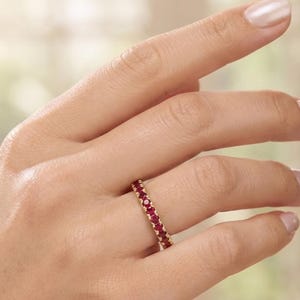 Könnte beinhalten: Ein goldener Eternity-Ring mit einer durchgehenden Reihe kleiner, runder, tiefroter Rubine in einer gemeinsamen Krappenfassung. Der Ring wird am Finger getragen und zeigt die leuchtend roten Steine auf dem polierten Goldband.