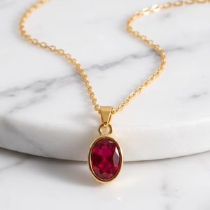 May include: A gold chain necklace featuring a single oval-shaped, faceted deep red gemstone pendant set in a smooth gold bezel. The delicate metal chain connects to the pendant via a small gold loop, creating a simple and elegant jewelry piece.
