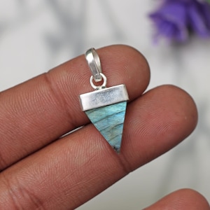 Puede incluir: Un colgante de plata con una piedra preciosa de labradorita triangular. La piedra preciosa es de color azul claro con destellos iridiscentes de verde y oro.