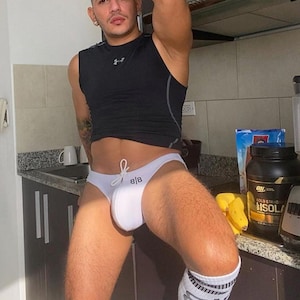 Könnte beinhalten: Ein schwarzes ärmelloses Kompressionsshirt und weiße Slips mit den Initialen "B|B" werden getragen. Die Person trägt auch weiße Socken mit schwarzen Akzenten. Eine Küchenzeile und einige Lebensmittel befinden sich im Hintergrund.