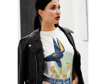 May include: A white t-shirt featuring a graphic of Anubis, the Egyptian god of the afterlife, in navy blue and gold. The shirt is paired with light blue jeans and a black leather jacket.