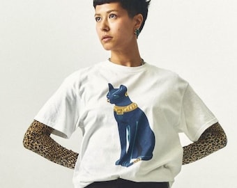 May include: A white t-shirt with a blue and gold illustration of a cat wearing a collar. The person wearing the shirt is also wearing leopard print arm warmers.