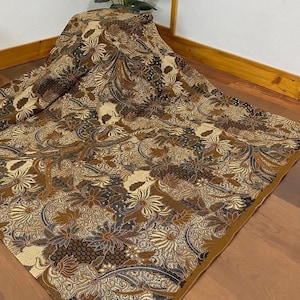 Puede incluir: Una tela con estampado marrón y dorado con un diseño floral. La tela está doblada y se muestra sobre una superficie de madera.