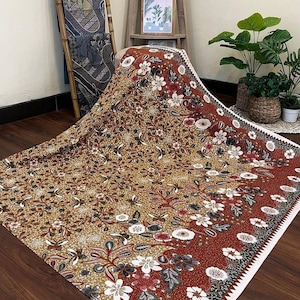 Puede incluir: Una tela grande, rectangular, de color marrón y rojo con estampado y ribete blanco. La tela tiene un diseño floral con flores blancas, rojas y negras. La tela está sobre un suelo de madera.