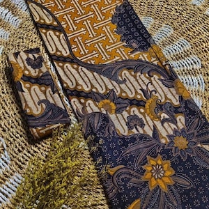 May include: Two pieces of fabric with a traditional batik pattern. One piece is a dark blue with a white and gold floral design. The other piece is a gold with a geometric pattern.