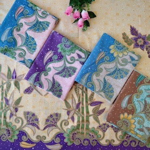 May include: Four folded fabric squares featuring intricate batik patterns in blue, purple, and brown, layered over a cream-colored textile base with bamboo and floral motifs. The fabric displays detailed botanical designs with green and pink accents.