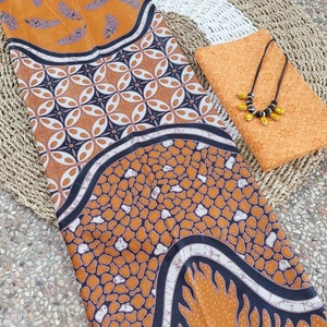 Può includere: Un tessuto batik marrone e nero con un motivo geometrico. Il tessuto è piegato a metà e ha un pezzo di tessuto più piccolo con un motivo simile e una collana di perline nere in cima.