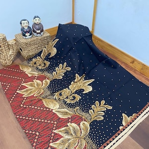Può includere: Tessuto nero con un motivo batik rosso e oro. Il tessuto ha un bordo bianco e piccoli punti dorati.