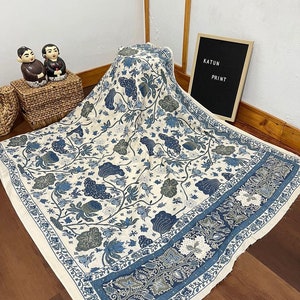 Può includere: Un tessuto bianco con un motivo floreale blu e verde. Il tessuto ha un bordo con un motivo floreale più intricato. Il testo "KATUN PRINT" è visibile su un cartello nero.