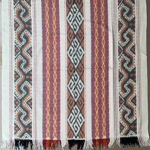 May include: A white fabric with black, orange, and red stripes. The stripes have a geometric pattern. The fabric has a fringe on the bottom.