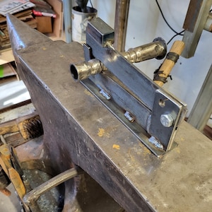 Puede incluir: Una herramienta de metal con un tubo conectado a ella está montada en un yunque grande. La herramienta está hecha de acero y tiene una forma rectangular. El yunque está hecho de hierro y tiene una parte superior redondeada.