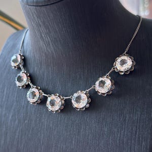 May include: A silver-toned necklace featuring seven round, clear faceted stones arranged in a row. Each stone is set within a scalloped metal frame, connected by thin silver-toned links that form a delicate chain around the neck area.