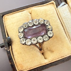 May include: An antique ring featuring a rectangular, lilac-coloured central stone, encircled by a halo of smaller, clear stones. The ring is set in a gold band and presented in a vintage, velvet-lined box with a dark exterior.