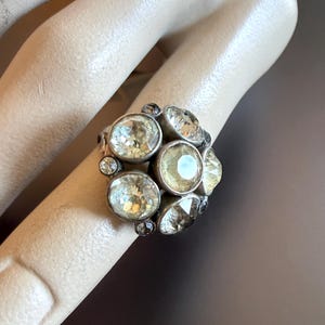Peut inclure: Une bague argentée ornée d'un groupe de cristaux ronds à facettes, de couleur jaune pâle et transparents, disposés en motif floral. La monture métallique présente une finition vieillie et sombre, avec des pierres de tailles variées créant un aspect texturé et multidimensionnel sur l'anneau.