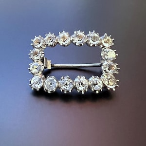 May include: A silver brooch with a rectangular frame set with clear rhinestones.