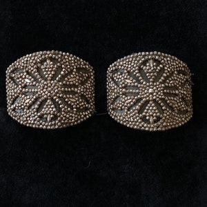May include: Two silver-toned metal embellishments with a starburst design. The embellishments are made of small beads and have a raised, textured surface.