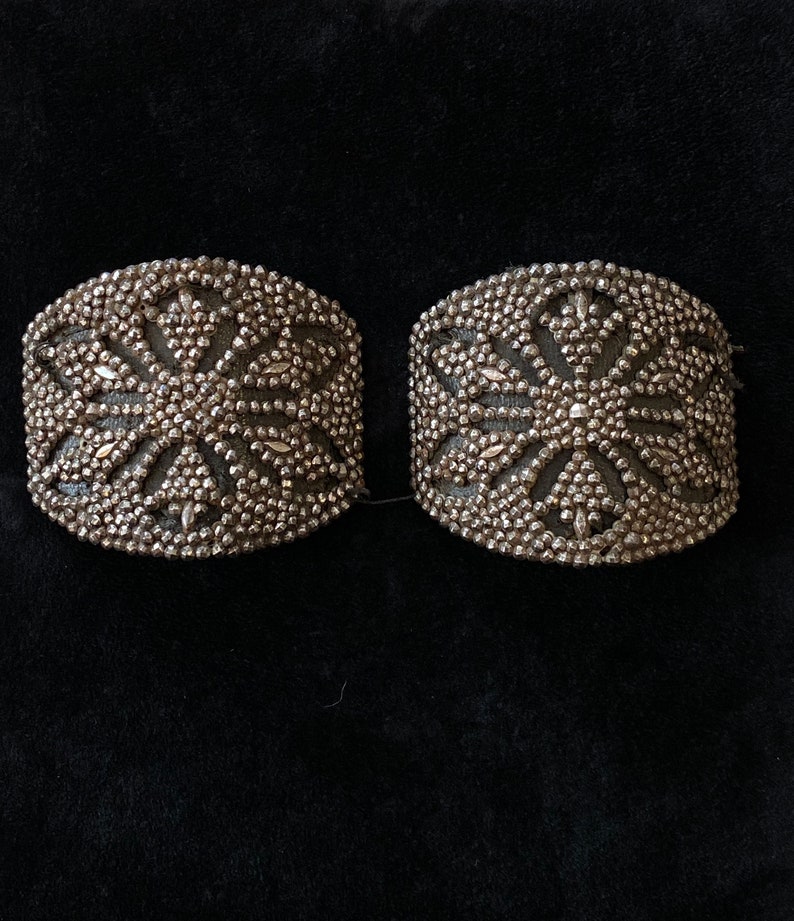 May include: Two silver-toned metal embellishments with a starburst design. The embellishments are made of small beads and have a raised, textured surface.