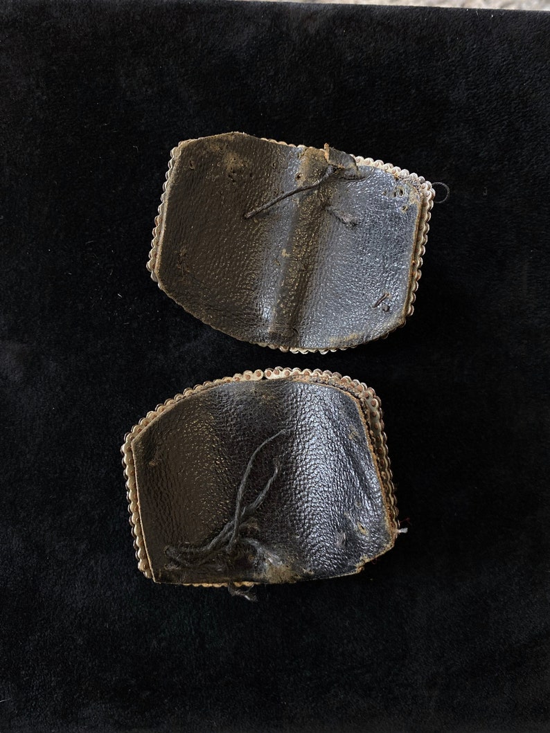 May include: Two black leather patches with silver beading around the edges. The patches are worn and have loose threads.