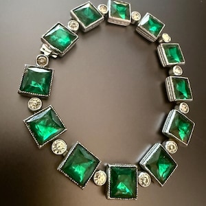 May include: A silver-toned bracelet featuring square, emerald-green gemstones set in a square frame. Small, round, clear gemstones are interspersed between the larger stones. The bracelet is displayed on a dark surface.