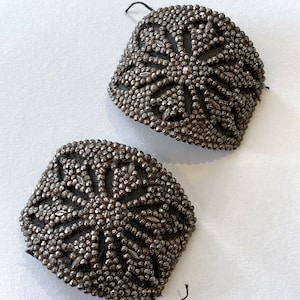 May include: Two silver metal embellishments with a starburst design. The embellishments are made of small metal beads and have a slightly curved shape.