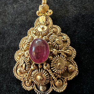 May include: A gold pendant with a pink gemstone set in the center. The pendant is intricately designed with a filigree pattern.