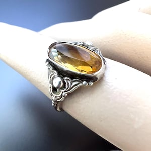 May include: A silver ring with an oval-shaped, faceted, amber-colored gemstone set in a filigree design.