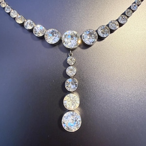 Peut inclure: Un collier composé d'une série de pierres rondes, transparentes et facettées, serties dans du métal sombre. Le design comprend une chaîne horizontale de pierres reliée à une section verticale composée de cinq pierres de tailles variées.