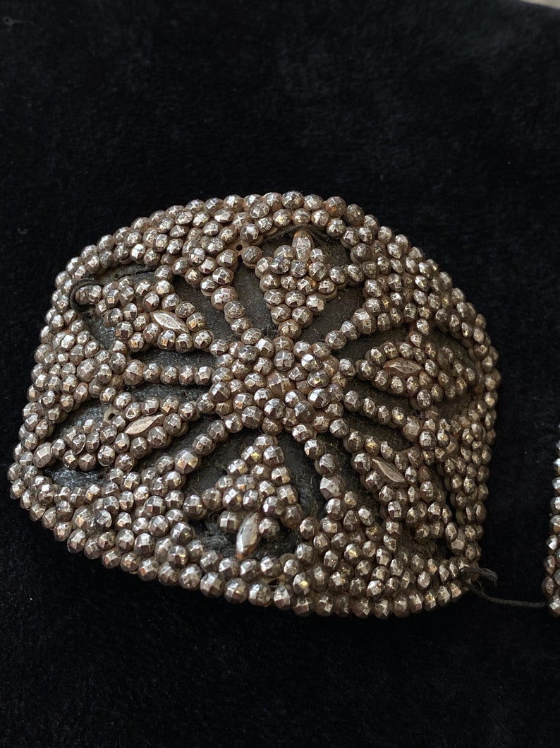 May include: A pair of silver-toned metal embellishments with a geometric design. The embellishments are made of small, round beads that are arranged in a pattern of overlapping circles.