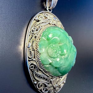 May include: A silver filigree pendant featuring a carved green jade stone. The jade stone is carved with a floral design. The pendant is on a silver chain.