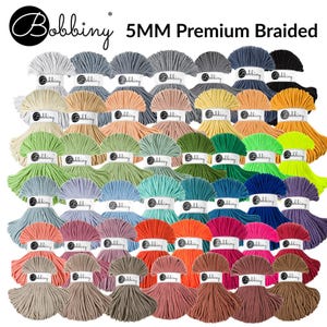 May include: A collection of Bobbiny 5MM Premium Braided yarn in various colors. The yarn is arranged in neat bundles, showcasing a wide spectrum of hues, including white, gray, green, blue, orange, pink, and brown. The Bobbiny logo is visible on each label.