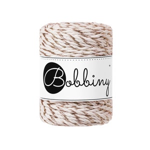 May include: A spool of off-white and gold twine with the word "Bobbiny" printed on a white label. The twine is tightly wound, and the gold strands add a subtle shimmer. Ideal for crafting and decorative projects.